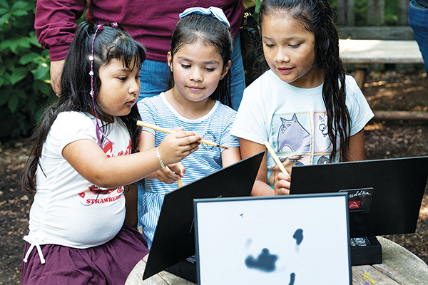 Three children are focused on painting with brushes on a white surface, surrounded by black palettes and art supplies outdoors.